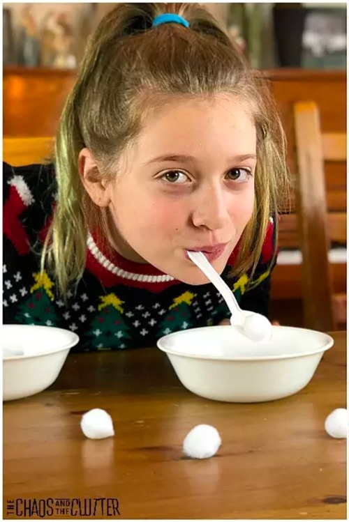 Girl in festive Christmas sweater playing the 'Snow Shovel Spoon Christmas Game' using her mouth to hold the spoon and scoop cotton balls into a bowl.