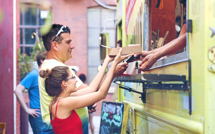 Woman taking a box of food at the window of a food truck while man pays for the food next to her