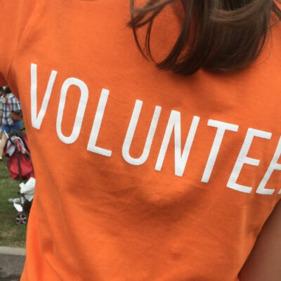 A close-up of a person wearing an orange t-shirt with the word 'VOLUNTEER' printed in large white letters on the back. The background shows a group of people outdoors, possibly participating in an event.