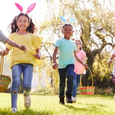Five children wearing colorful bunny ears headbands run across a grassy field carrying Easter baskets, smiling and laughing. The bright outdoor setting with trees and sunlight suggests a fun Easter egg hunt, ideal for church or community events.